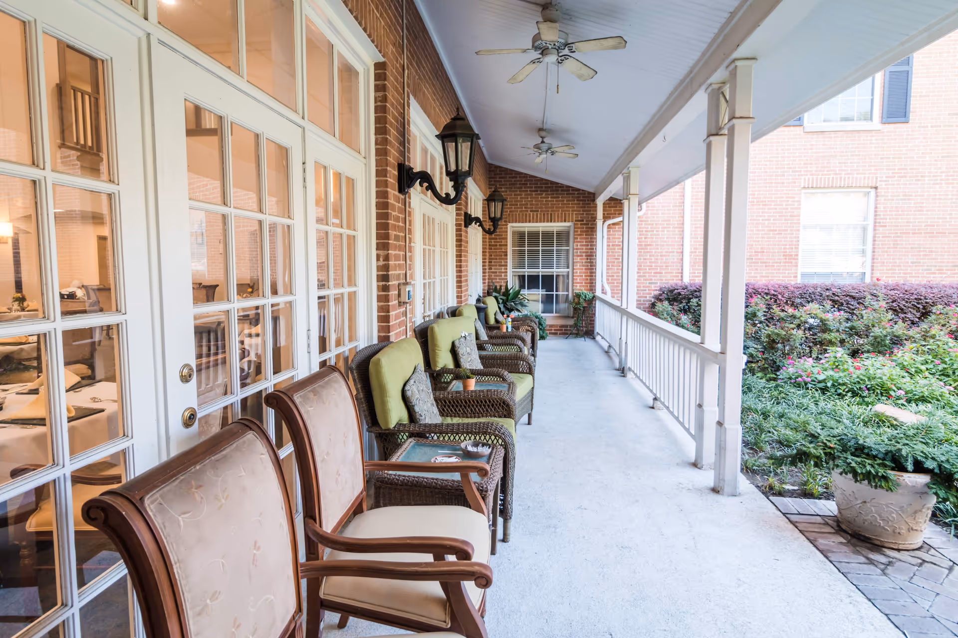 Covered outdoor porch area with several cushioned wicker chairs and wooden armchairs arranged along a brick wall with white-framed glass doors. Ceiling fans and wall-mounted lantern-style lights are visible overhead. On the right side, there is a white railing overlooking a garden with green shrubs and flowering plants.