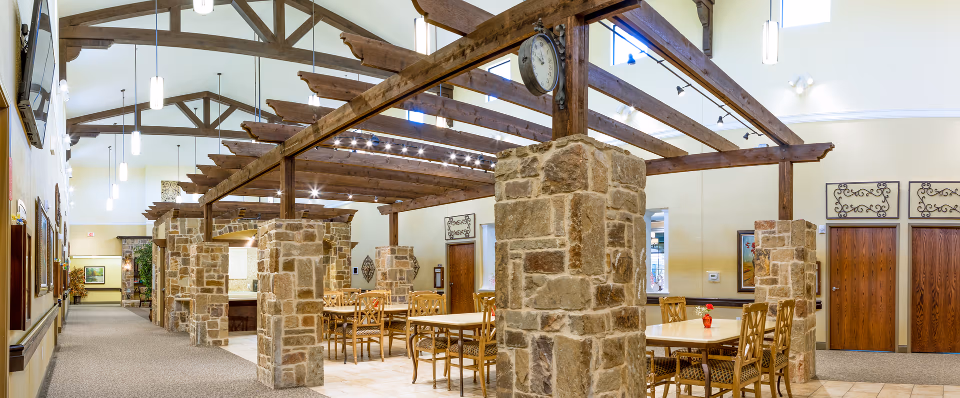 Interior view of a spacious dining area with wooden beams and stone pillars. Several tables with chairs are arranged neatly, and pendant lights hang from the ceiling. The room has a warm and inviting atmosphere with decorative wall art and a clock mounted on one of the stone pillars.