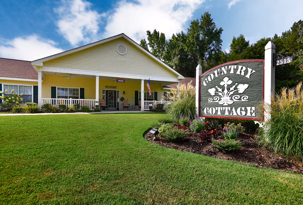 Exterior view of Country Cottage Decatur, a single-story yellow building with a covered porch, white railings, and green shutters. The front lawn is well-maintained with green grass and landscaped flower beds. A large wooden sign with the words 'Country Cottage' is prominently displayed near the entrance, along with the street number 2515. Trees and a partly cloudy blue sky are visible in the background.