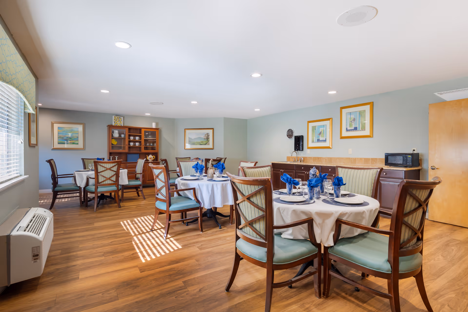 Bright dining room with round tables set with blue napkins and wooden chairs in a senior living facility.