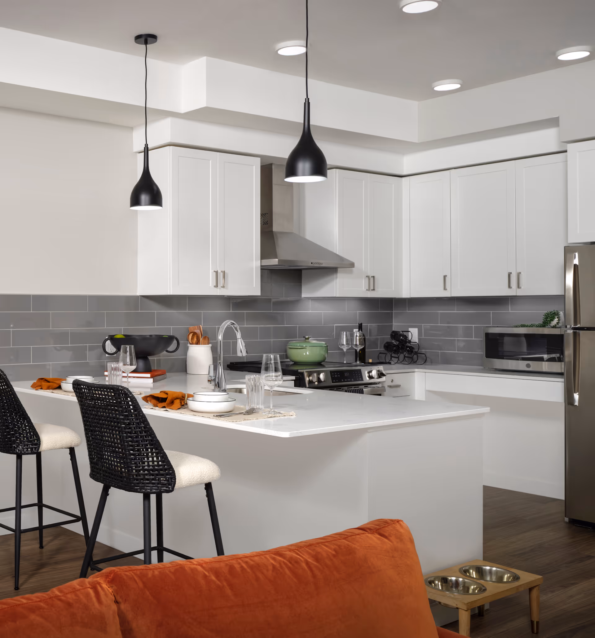 Modern kitchen with white cabinets, gray subway tile backsplash, stainless steel appliances, and a white island countertop with two black woven bar stools. Two black pendant lights hang above the island. An orange couch is partially visible in the foreground with a wooden pet feeding station on the floor nearby.