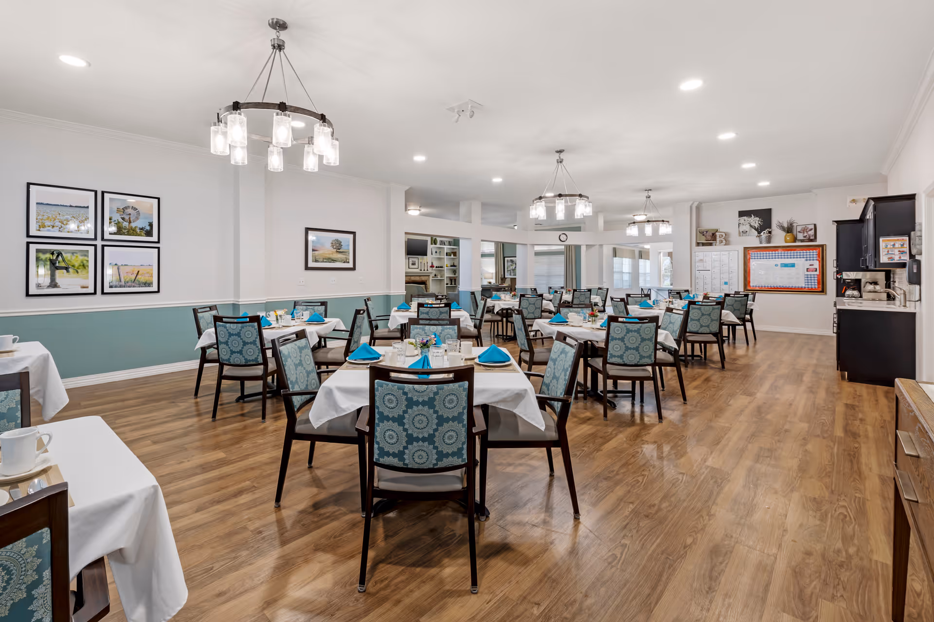 A spacious dining room with multiple tables covered in white tablecloths and set with blue napkins. The chairs have patterned blue upholstery. The room has wooden flooring, white walls with teal wainscoting, and several framed pictures on the walls. Modern chandeliers hang from the ceiling, and there is a cabinet and bulletin board visible in the background.