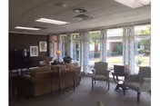 Sunlit communal lounge with sofas, armchairs, small tables, a television, and large windows with patterned curtains overlooking a courtyard.