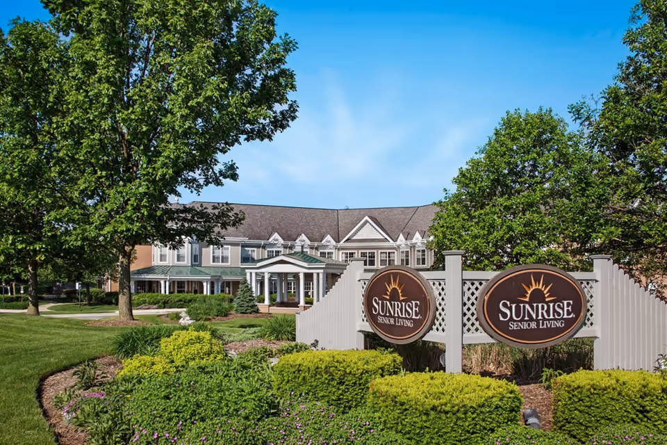 Exterior view of Sunrise Senior Living facility with a large sign in front surrounded by well-maintained landscaping, trees, and green grass under a clear blue sky.