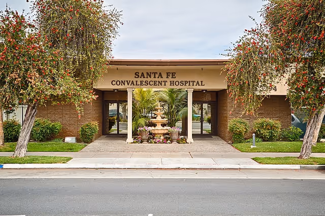 Front exterior view of Santa Fe Convalescent Hospital building with two trees on either side and a decorative fountain with plants in the entrance area.