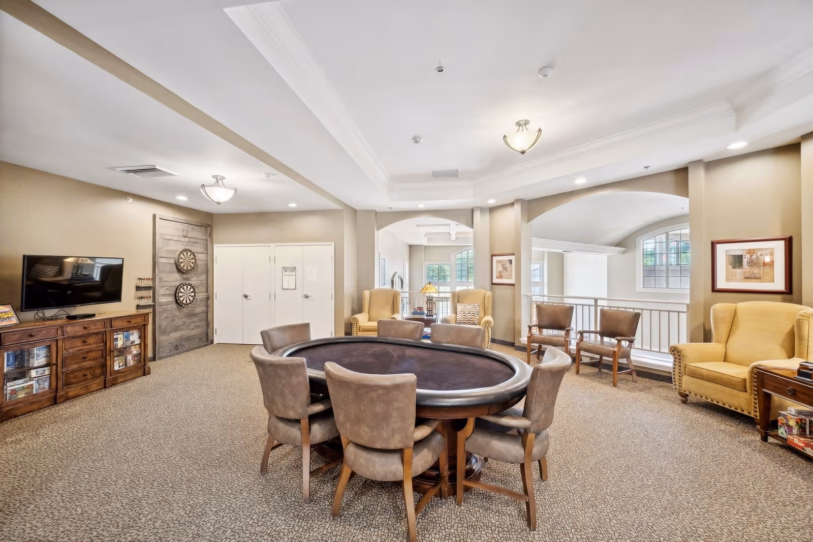A spacious common area with a round poker table surrounded by eight chairs in the center. The room features beige walls and carpeted floors. There are two dartboards mounted on a wooden panel near a flat-screen TV on a wooden cabinet. Comfortable armchairs and additional seating are arranged near large windows that let in natural light. The ceiling has recessed lighting and decorative light fixtures.