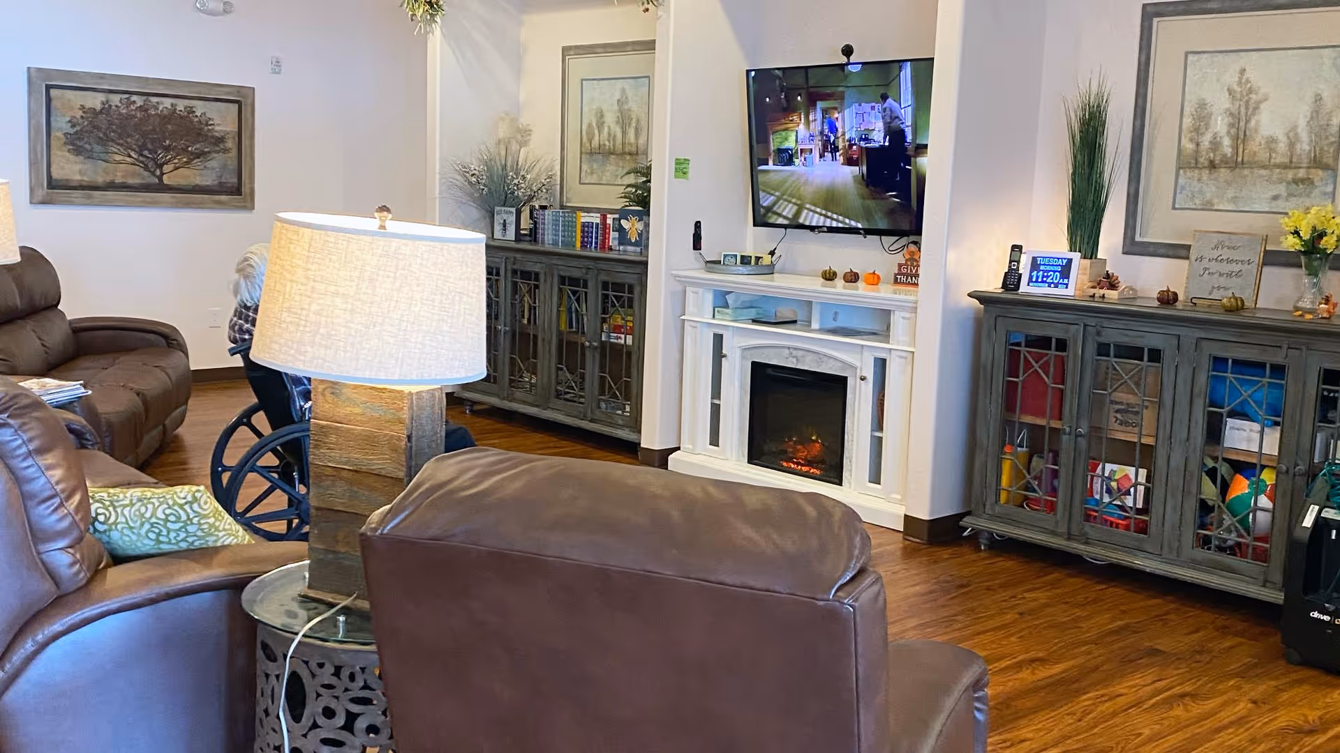 Cozy common living area with brown leather recliners, a side lamp, a wall-mounted TV above an electric fireplace, and storage cabinets.