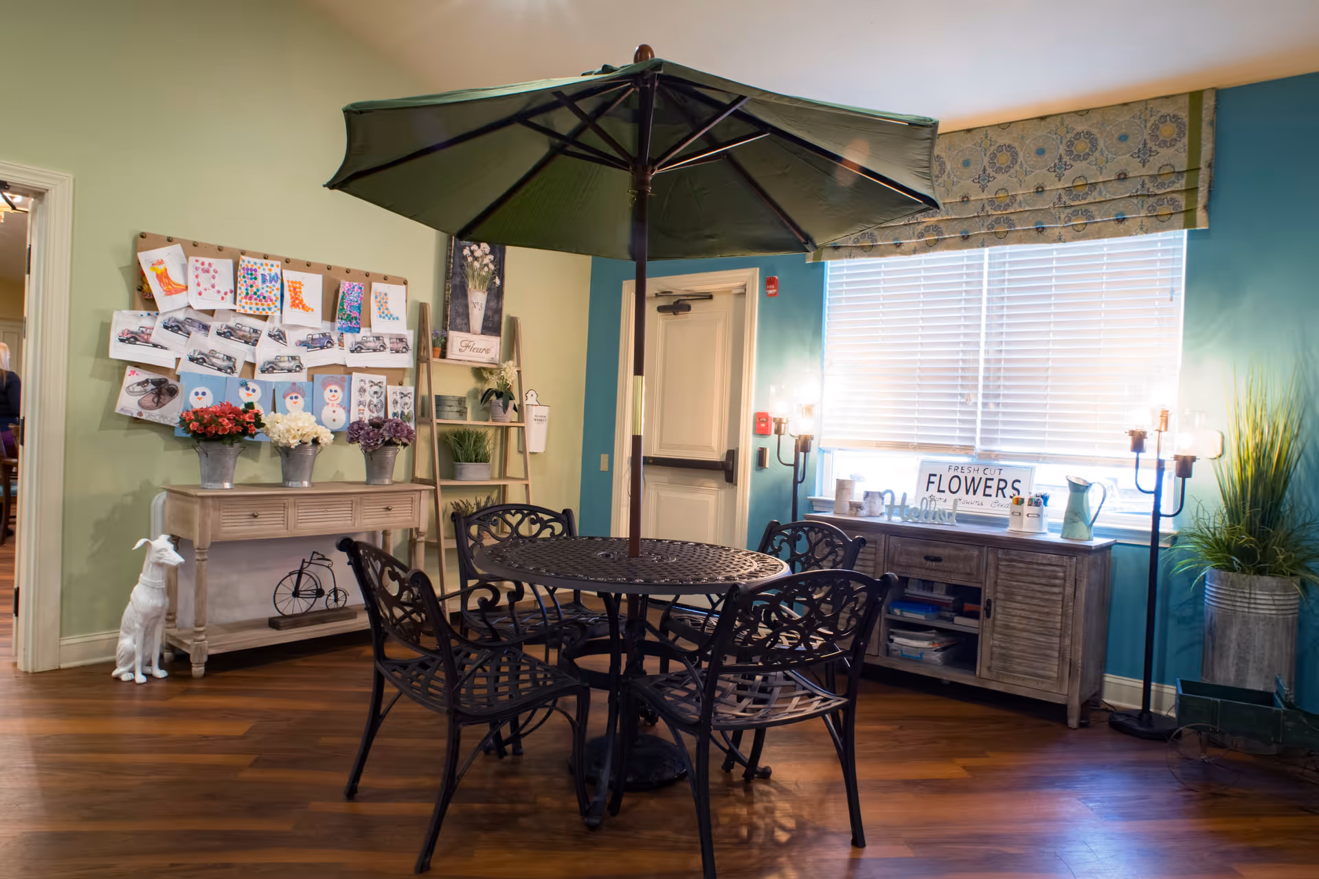 Indoor seating area with a round black metal table and four matching chairs under a large green umbrella. The room has wooden flooring, light green and teal walls, and a window with white blinds and a patterned valance. There is a wooden sideboard with decorative items and a sign that reads 'FRESH CUT FLOWERS'. On the left wall, there is a bulletin board with colorful artwork and a small table with flower pots. A white dog statue is placed on the floor near the table.