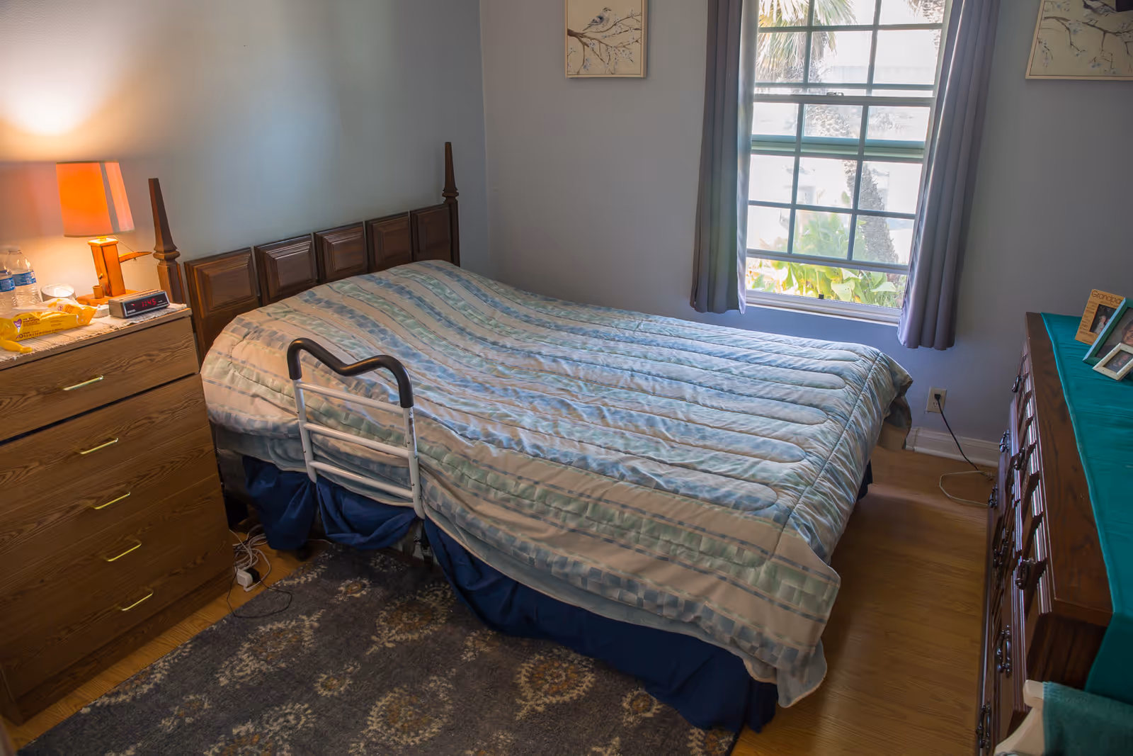 A bedroom with a bed covered in a striped comforter, a wooden headboard, and a bed assist rail. There is a wooden dresser with a lamp, water bottles, and snacks on the left side. On the right side, there is another wooden dresser with framed photos and a green cloth on top. A window with gray curtains lets in natural light, and a patterned rug is on the wooden floor.