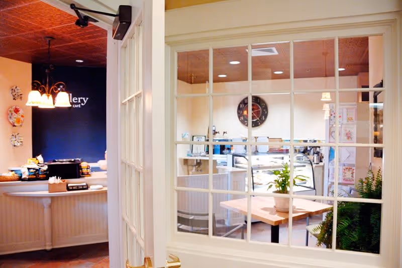 View through a glass-paneled door into a cozy café area with a wooden table, potted plants, and a clock on the wall. To the left, a counter with snacks and a hanging light fixture is visible, with the word 'Gallery' partially seen on a dark wall.