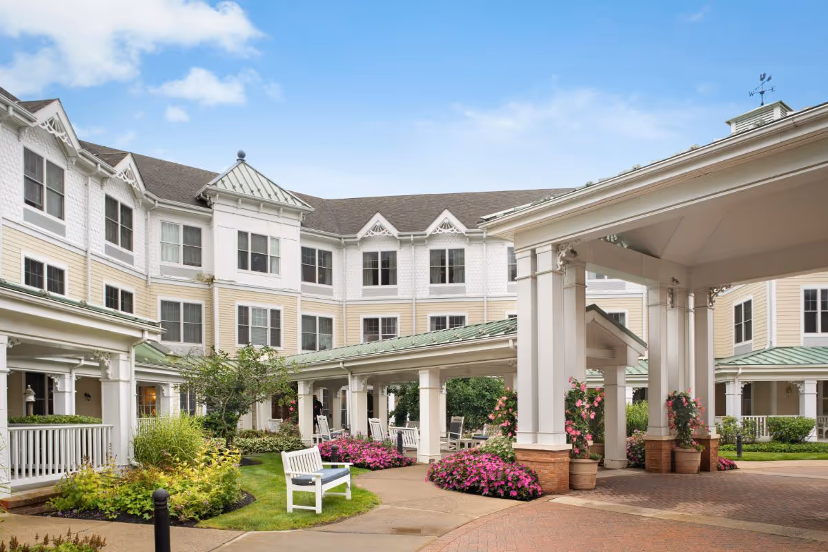 Covered entrance and landscaped courtyard of a multi-story beige-and-white senior living building with benches and flowerbeds.