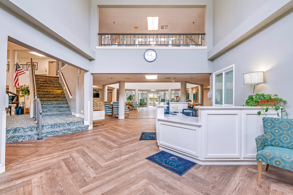 Bright and spacious senior living facility lobby with a white reception desk on the right, a patterned blue chair, a potted plant, and a lamp on the wall. The floor has a wood pattern, and there is a staircase with a carpeted runner on the left. The area beyond the reception desk has seating with chairs and tables, and a clock is mounted on the wall above an open balcony area.
