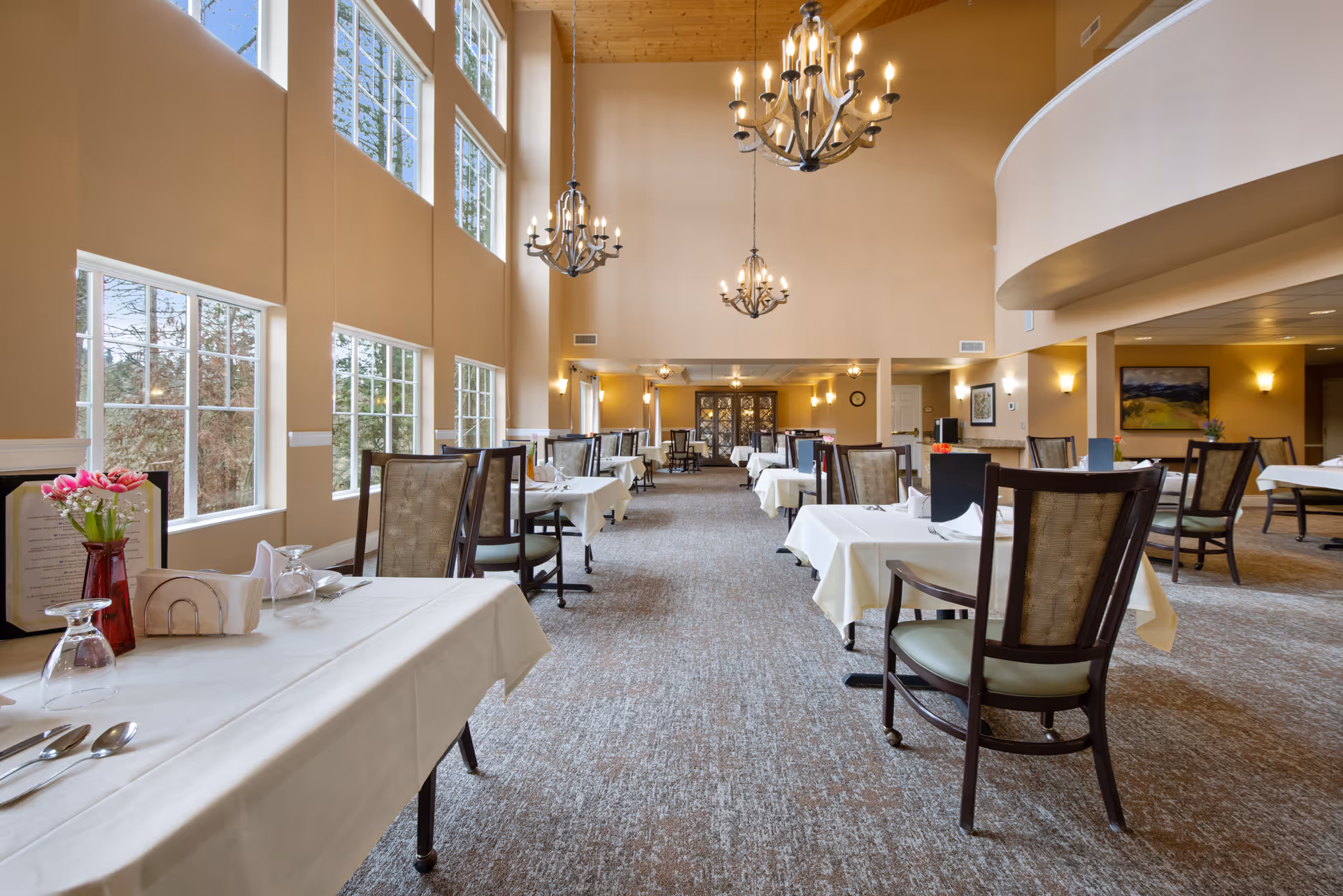 A spacious dining room with high ceilings and large windows letting in natural light. Tables are covered with white tablecloths and set with utensils, glasses, and napkins. There are chandeliers hanging from the ceiling and chairs arranged neatly around the tables. The room has a warm, inviting atmosphere with beige walls and carpeted floors.