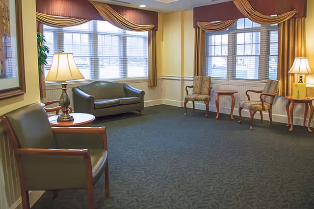 A sitting area in a senior living facility with green upholstered chairs and a matching loveseat arranged around wooden side tables with lamps. The room has large windows with brown and gold curtains, beige walls with white wainscoting, and a dark patterned carpet.