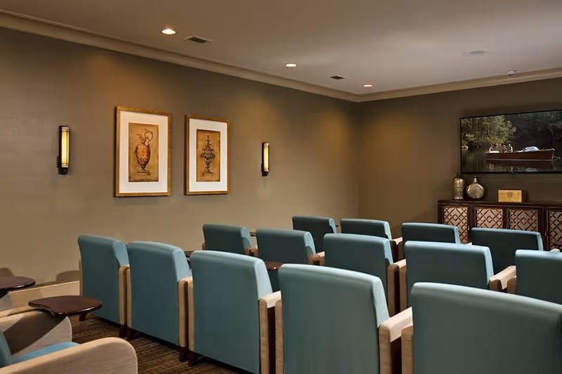 A small theater or media room with rows of teal upholstered chairs facing a large flat-screen TV mounted on the wall. The walls are painted beige and decorated with two framed artworks and wall sconces. A wooden cabinet with decorative items is below the TV.