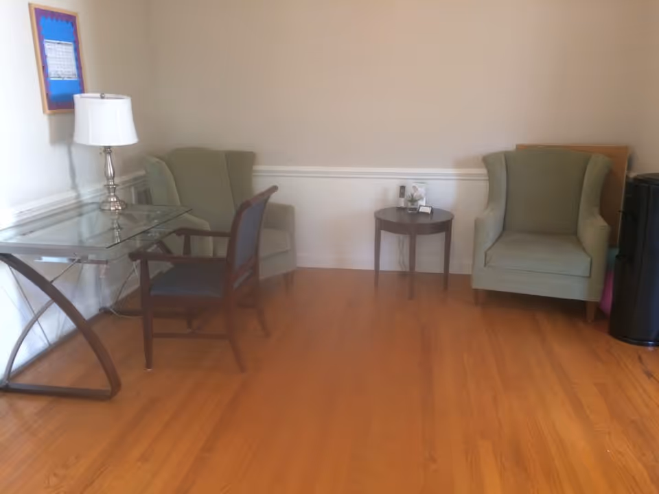 A small sitting area with two green upholstered armchairs, a wooden chair with a dark cushion, a glass-top desk with a silver lamp, and a round wooden side table with a phone and a small decorative item. The room has light-colored walls and wooden flooring.