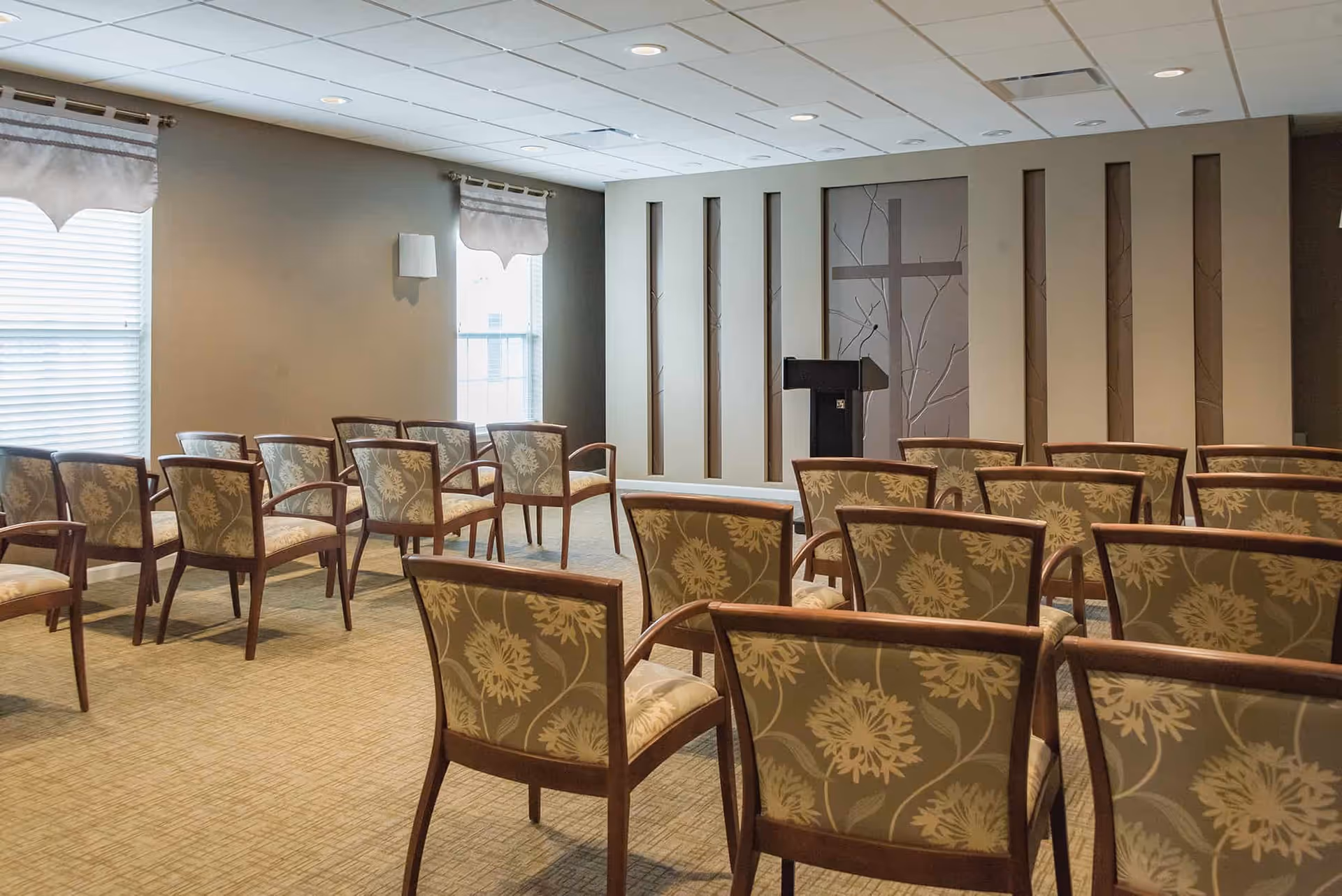 Rows of upholstered chairs facing a podium and decorative wall panels in a small meeting/chapel room.