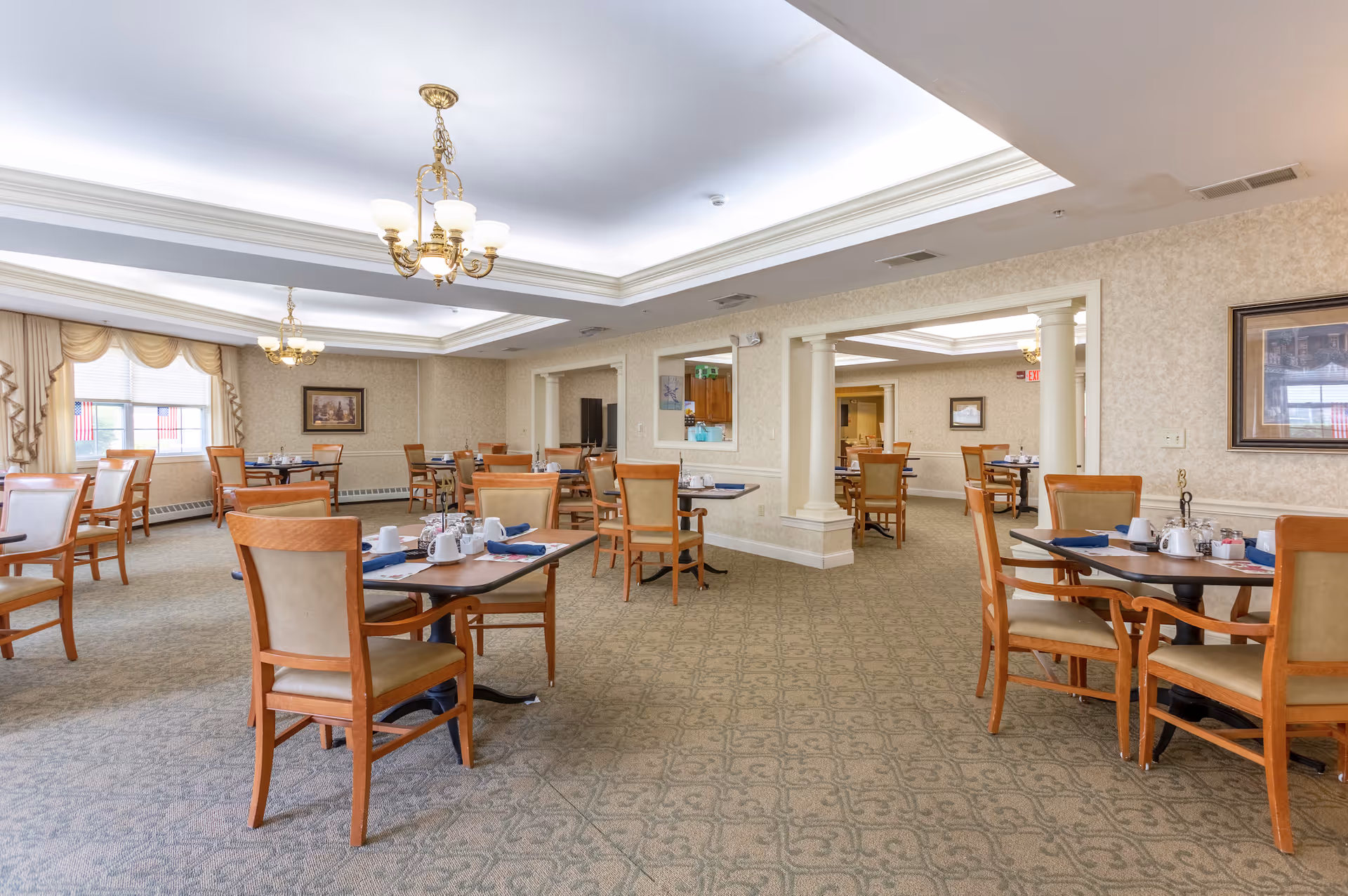 A spacious dining room with multiple wooden tables and chairs arranged neatly. Each table is set with white cups, napkins, and condiments. The room has beige patterned carpet, cream-colored walls with framed artwork, and large windows with draped curtains allowing natural light. Elegant chandeliers hang from the ceiling providing warm lighting.