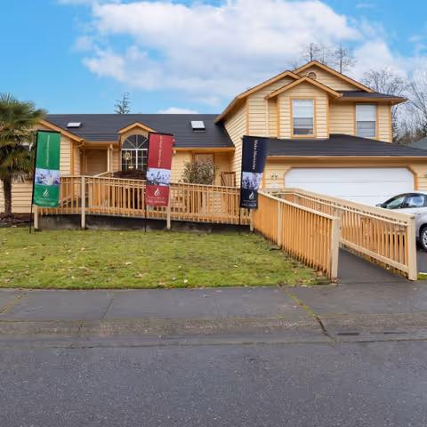 Exterior view of a two-story beige house with a wooden wheelchair ramp leading to the front entrance. There are three vertical banners on poles in front of the house, and a silver car is parked in the driveway. The sky is partly cloudy.