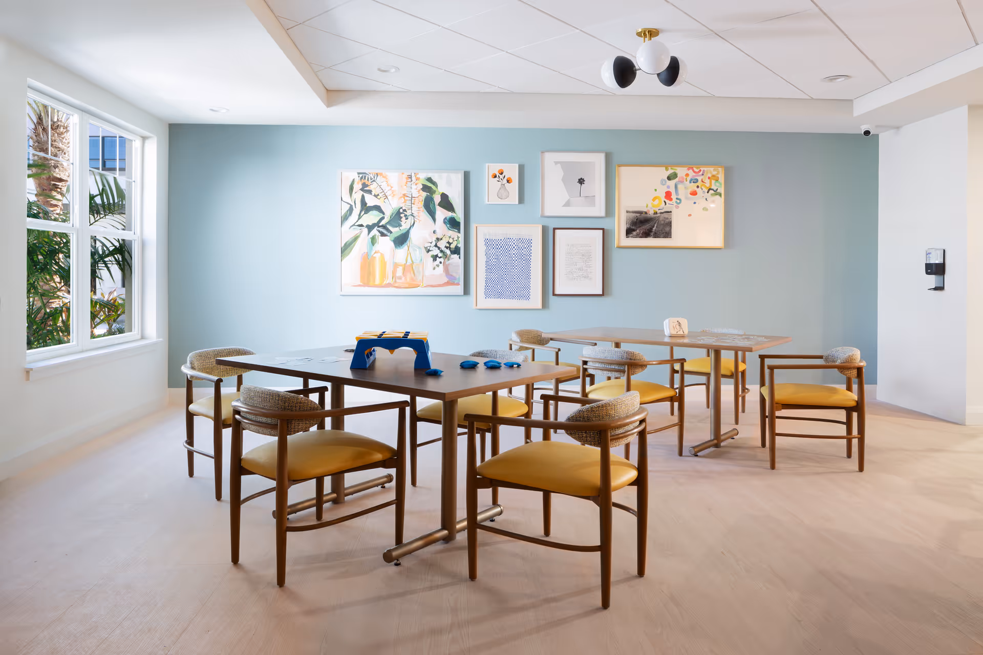 A bright, modern room with light wood flooring and a light blue accent wall adorned with six framed artworks. The room contains two wooden tables with yellow cushioned chairs arranged around them. A large window on the left side lets in natural light and shows greenery outside. The ceiling has a white grid pattern with a modern black and white light fixture. A hand sanitizer dispenser is mounted on the right wall.