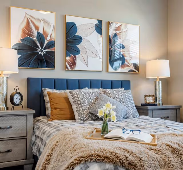 A cozy bedroom featuring a bed with a blue upholstered headboard, multiple decorative pillows, and a beige furry blanket. On the bed is a wooden tray holding a small vase with yellow flowers and an open book with blue reading glasses. On each side of the bed are matching wooden nightstands with table lamps, an alarm clock on the left nightstand, and a framed photo on the right nightstand. Above the bed are three framed floral artworks with blue and beige tones.