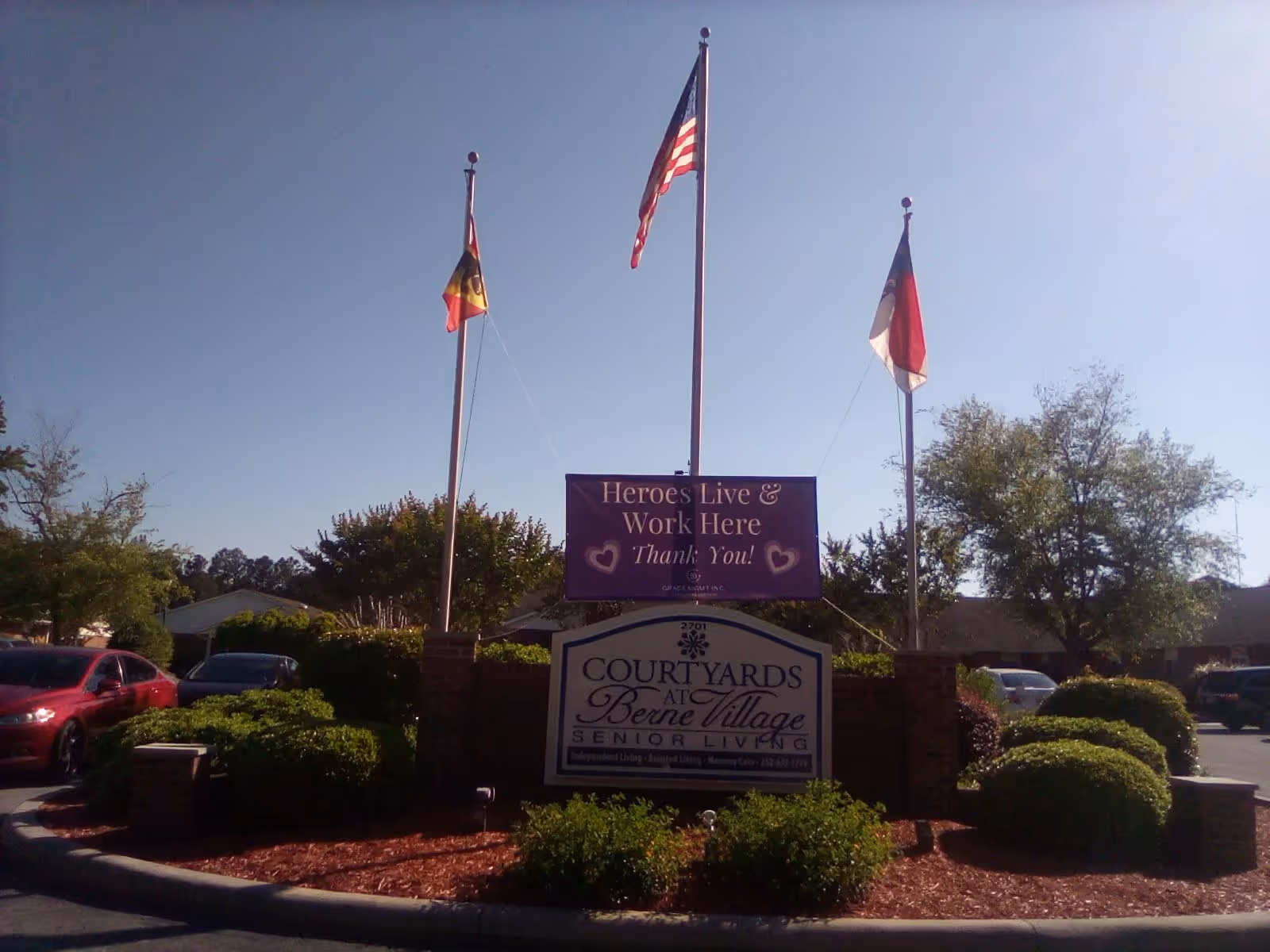 Entrance sign for Courtyards at Berne Village Senior Living with three flagpoles displaying flags behind it. A purple banner above the sign reads 'Heroes Live & Work Here Thank You!' There are cars parked and trees in the background under a clear sky.