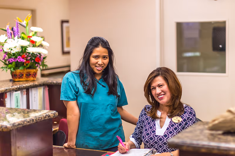 Two women smiling at a reception desk in a nursing and rehabilitation center. One woman is standing and wearing teal scrubs, while the other is sitting and writing in a book, wearing a patterned blouse with a name badge. There is a flower arrangement on the counter and a window in the background.