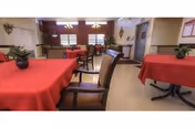 Interior view of a dining room with tables covered in red tablecloths, chairs arranged around the tables, potted plants as centerpieces, and windows letting in natural light.