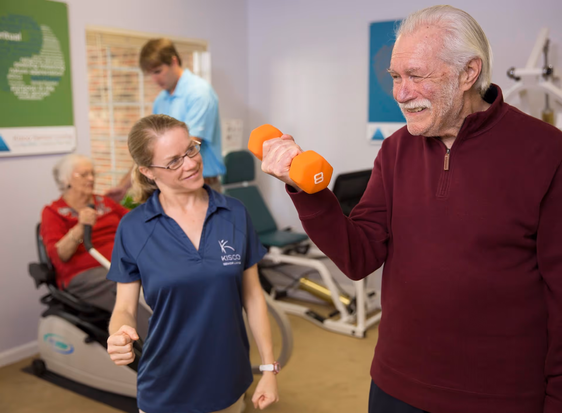 An elderly man in a maroon sweater is lifting an orange dumbbell while smiling. A woman in a blue KISCO shirt is standing next to him, encouraging him. In the background, an elderly woman is using a stationary exercise bike and a man in a light blue shirt is standing near exercise equipment in a fitness room.