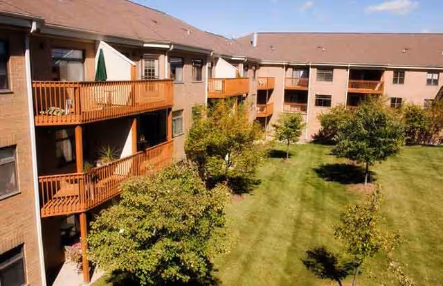 Exterior view of a multi-story residential building with balconies overlooking a well-maintained grassy courtyard with several small trees under a clear blue sky.