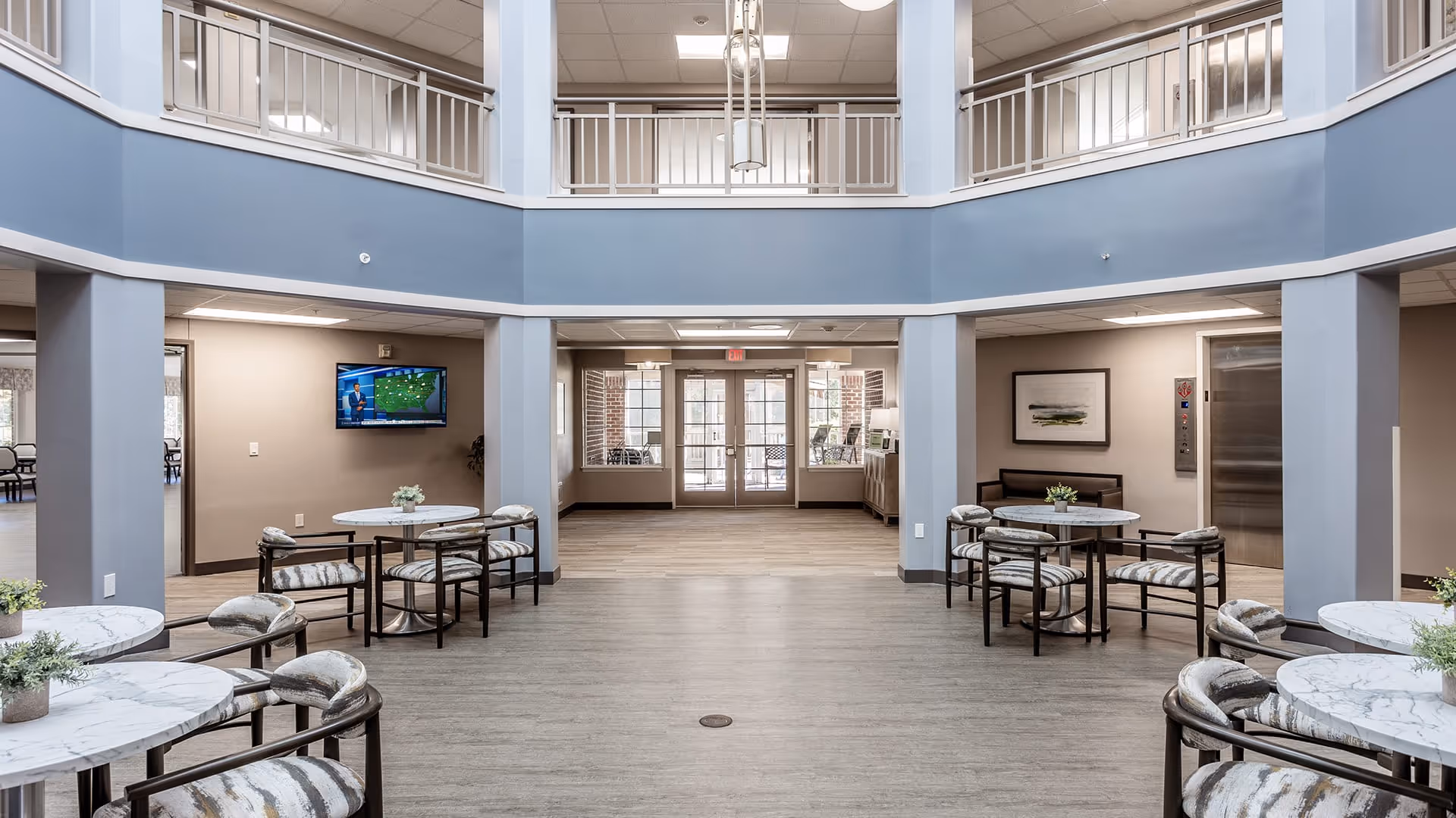 A spacious common area in a senior living facility with round tables and cushioned chairs arranged around the room. The walls are painted in neutral tones with a blue upper section and white railings on the second floor balcony. There is a television mounted on the left wall, an elevator on the right, and glass doors leading outside at the far end. Potted plants decorate the tables and the space has a bright, welcoming atmosphere.