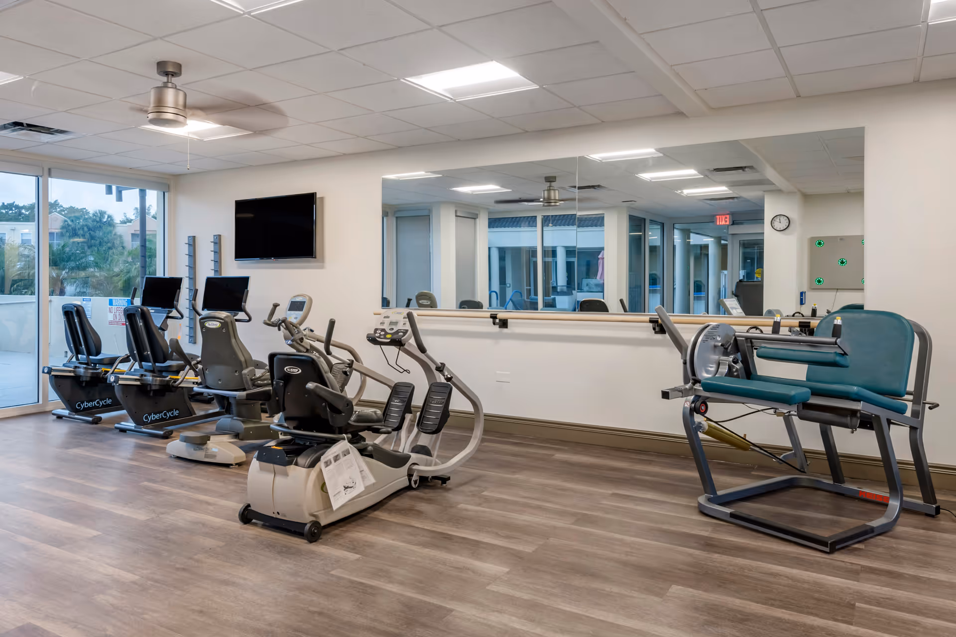 A fitness room with exercise equipment including recumbent stationary bikes and a leg press machine. The room has wood flooring, a large mirror on one wall, a ceiling fan, a wall-mounted TV, and large windows letting in natural light.