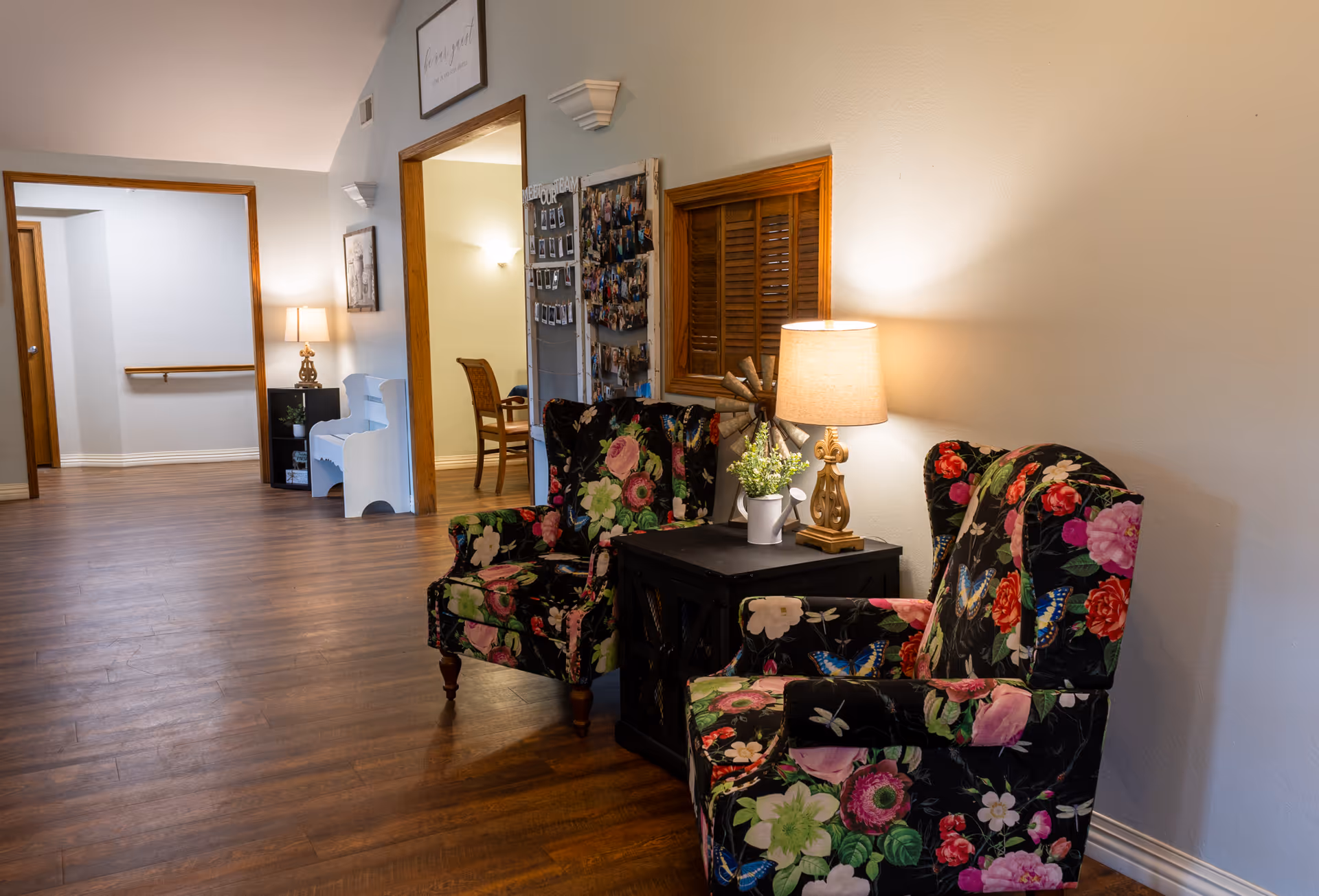 A cozy interior corner of a senior living facility with two floral-patterned armchairs facing each other and a small black side table between them. On the table is a lamp with a beige shade and a small white watering can with green plants. The room has wooden flooring and light-colored walls. In the background, there is a hallway with a white piano bench, a lamp on a small table, and a bulletin board with photos on the wall.