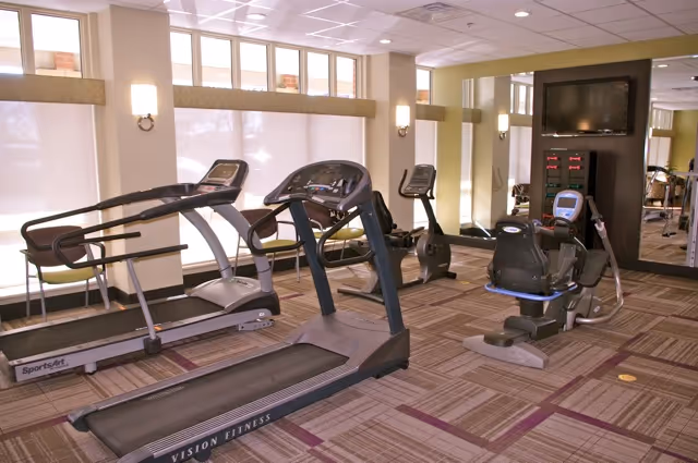 Indoor fitness room with exercise equipment including treadmills, stationary bikes, and a recumbent bike. The room has large windows with blinds, wall-mounted lights, and a wall-mounted TV above a control panel.