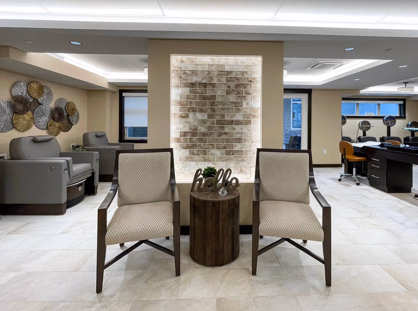 Bright communal lounge with two upholstered chairs flanking a small round table in front of a backlit decorative brick feature wall.