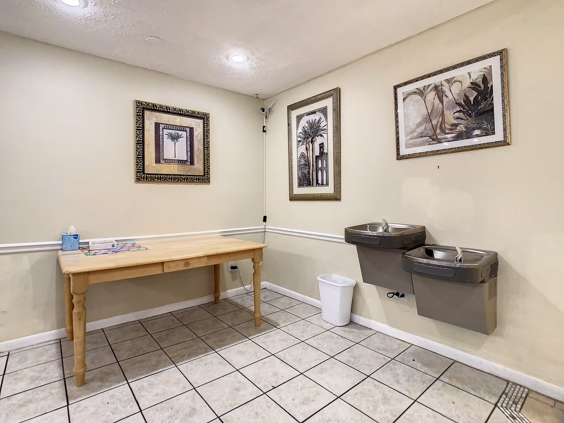 A small room with beige walls and tiled floor featuring a wooden table with tissues and a box on it, two wall-mounted drinking fountains, a white trash bin, and three framed pictures of palm trees on the walls.