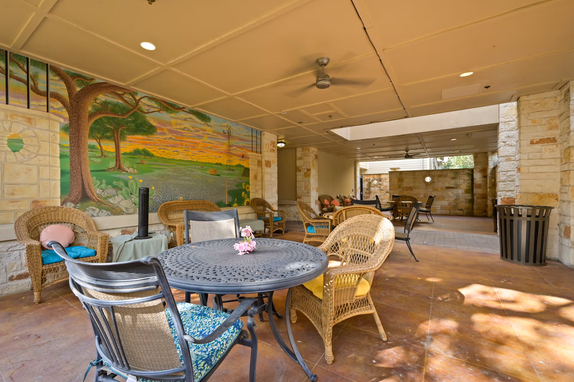 Covered outdoor seating area with wicker chairs and metal tables, featuring a colorful mural of a tree and landscape on one wall. The space has a ceiling fan, stone pillars, and a trash can, with sunlight casting shadows on the floor.