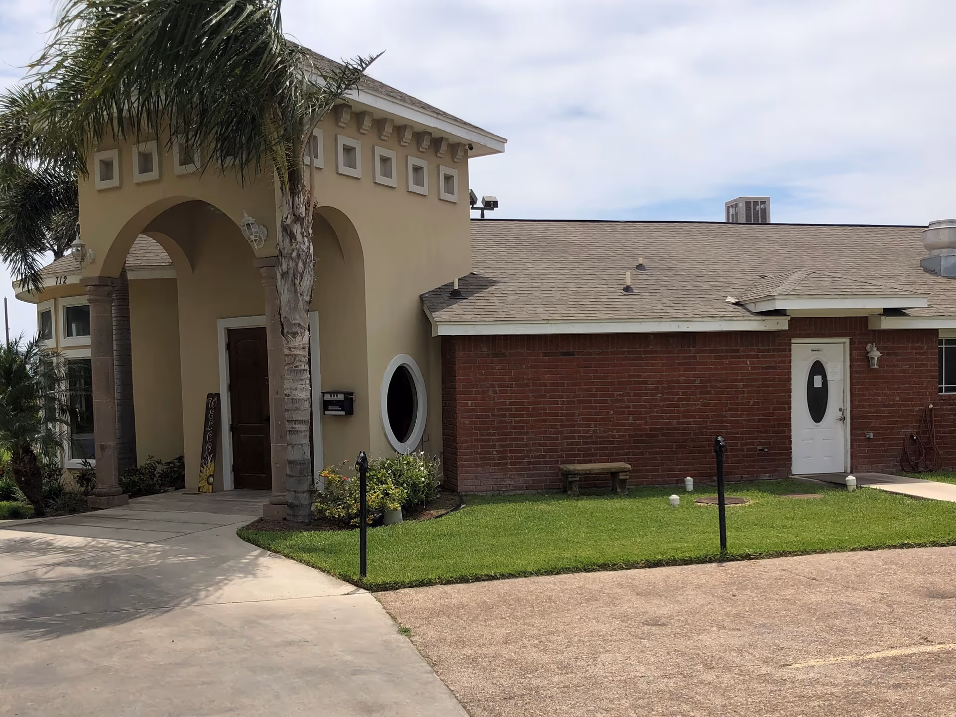 Exterior view of a building with a beige and brick facade, featuring an arched entrance supported by columns, a palm tree, a small green lawn, and a concrete driveway. The sky is partly cloudy.