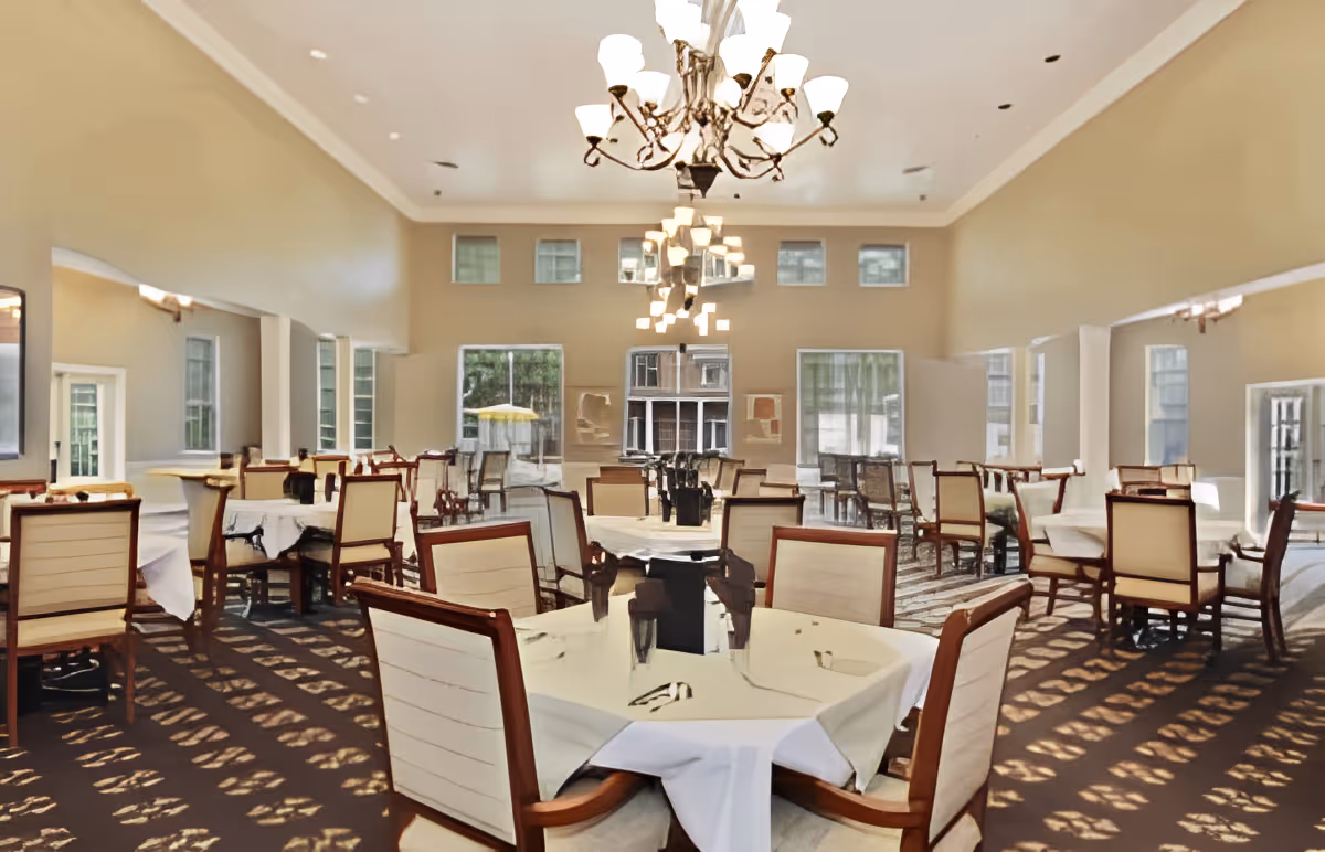 A spacious dining room with multiple tables covered in white tablecloths and surrounded by wooden chairs with cream-colored cushions. Large windows allow natural light to fill the room, and elegant chandeliers hang from the ceiling. The room has beige walls and a patterned carpet.