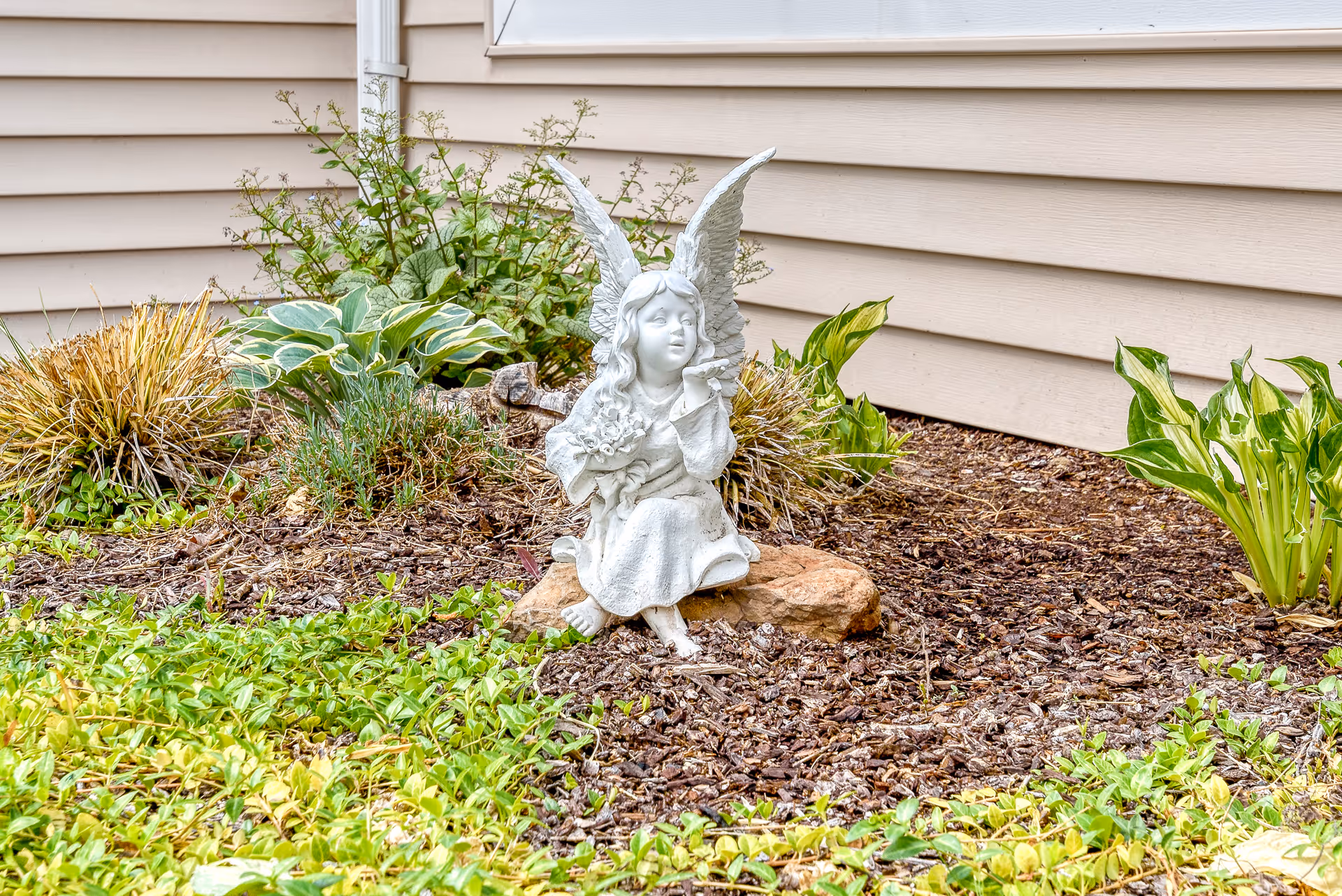 Small angel statue with wings seated in a mulched garden bed beside beige house siding.