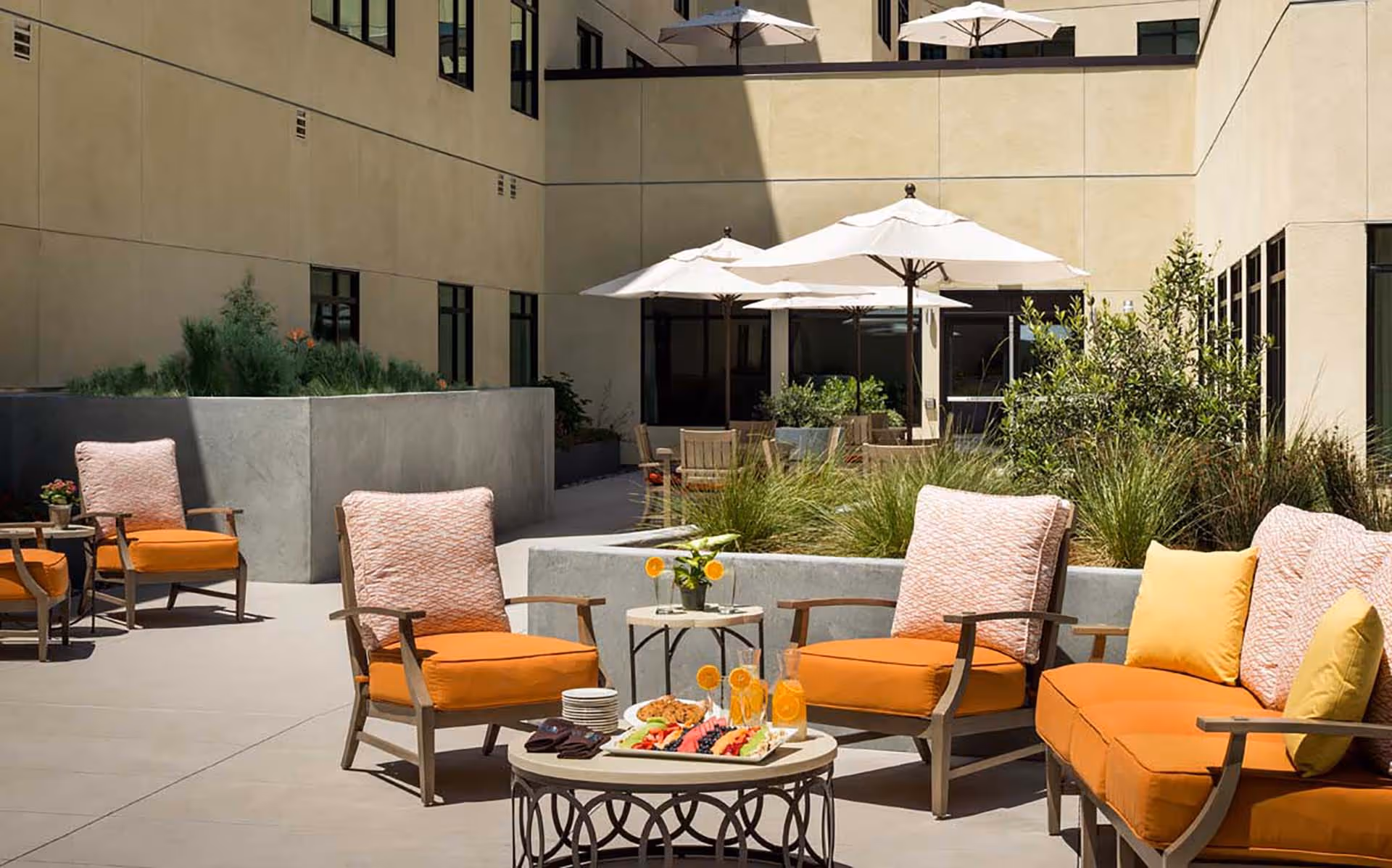 Outdoor patio area with orange cushioned chairs and a sofa, a small table with plates, drinks, and snacks, surrounded by planters with greenery and beige building walls with windows in the background.