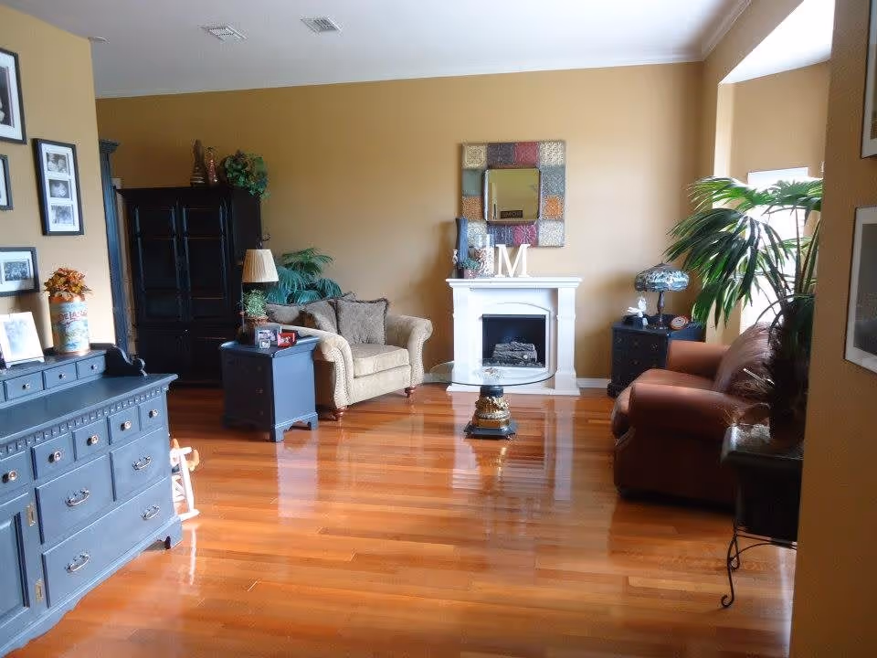 A cozy living room with polished wooden floors, beige and brown armchairs, a white fireplace with decorative items on the mantel, a colorful framed mirror above the fireplace, black side tables with lamps, green plants, and framed pictures on the walls.