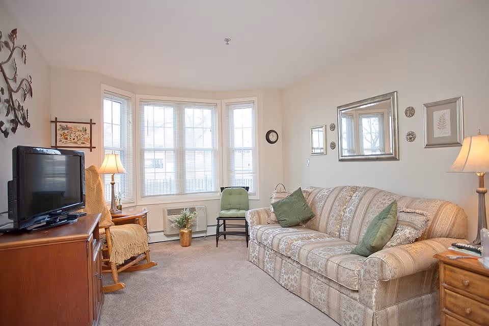 A cozy living room with a patterned beige sofa adorned with green pillows, a wooden rocking chair with a yellow throw, a wooden TV stand with a flat-screen TV, a small green chair near a bay window with blinds, and two table lamps providing warm lighting. The walls are decorated with framed art and mirrors.