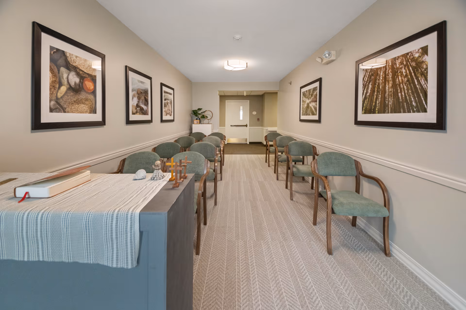 A hallway with two rows of green cushioned chairs along both walls. The walls are decorated with framed nature photographs. At the end of the hallway is a closed door with a small window. In the foreground, there is a table covered with a striped cloth, holding a book and small decorative items including crosses and a figurine.