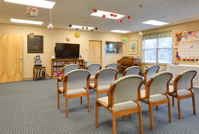 A community activity room with rows of wooden-framed chairs facing a TV and bookshelf, a piano by the window, and hanging ceiling decorations.