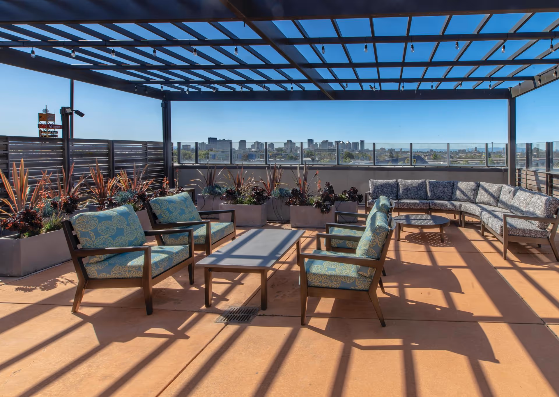 Outdoor patio area with cushioned chairs and a curved sofa under a pergola, overlooking a city skyline with clear blue skies. Planters with various plants line the perimeter.