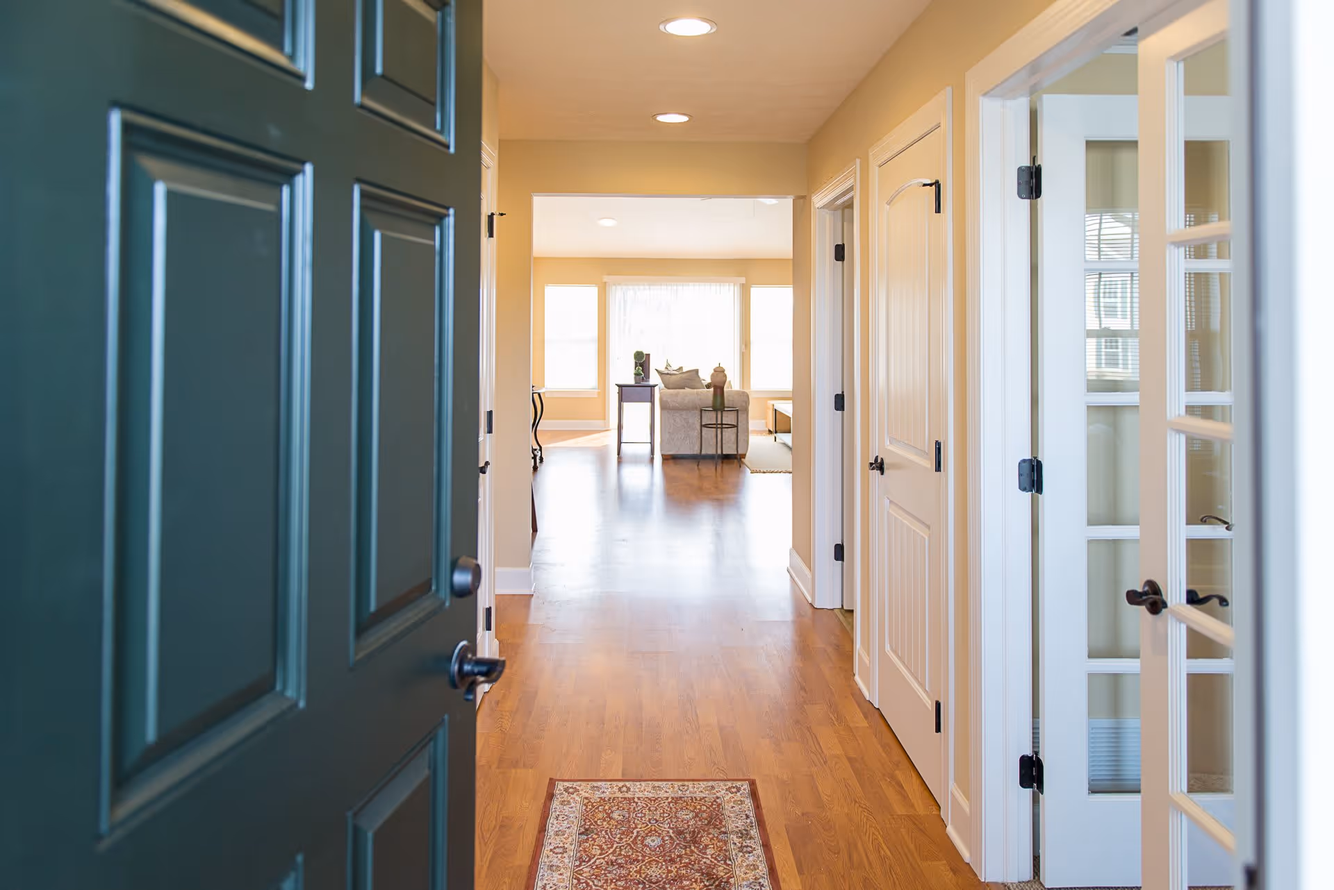 View through an open dark green front door into a hallway with wooden flooring and beige walls. The hallway leads to a bright living room with large windows, a sofa, and side tables. There are several white doors along the hallway, including a glass-paneled door on the right.