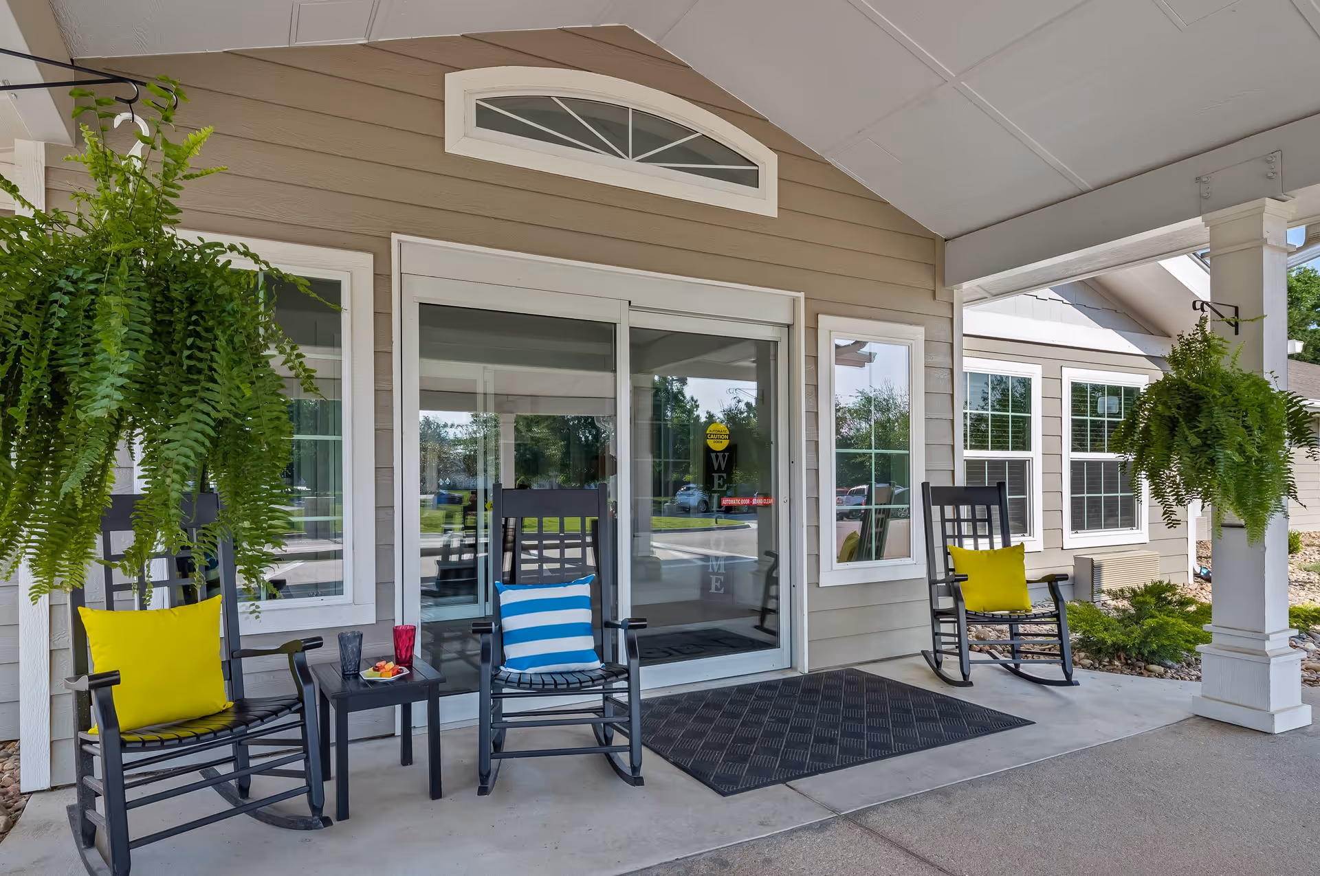 Front entrance of a senior living facility with a covered porch featuring three black rocking chairs with colorful cushions, two hanging green ferns, a small table with drinks and snacks, and glass sliding doors with a welcome sign.