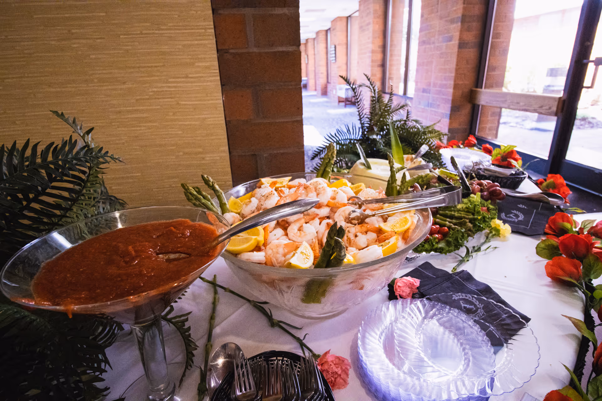 A buffet table set up with a large bowl of shrimp garnished with lemon wedges and asparagus, accompanied by a large glass bowl of cocktail sauce. The table is decorated with green plants and red flowers, with clear plastic plates and utensils nearby. The setting appears to be indoors near large windows with a brick wall in the background.