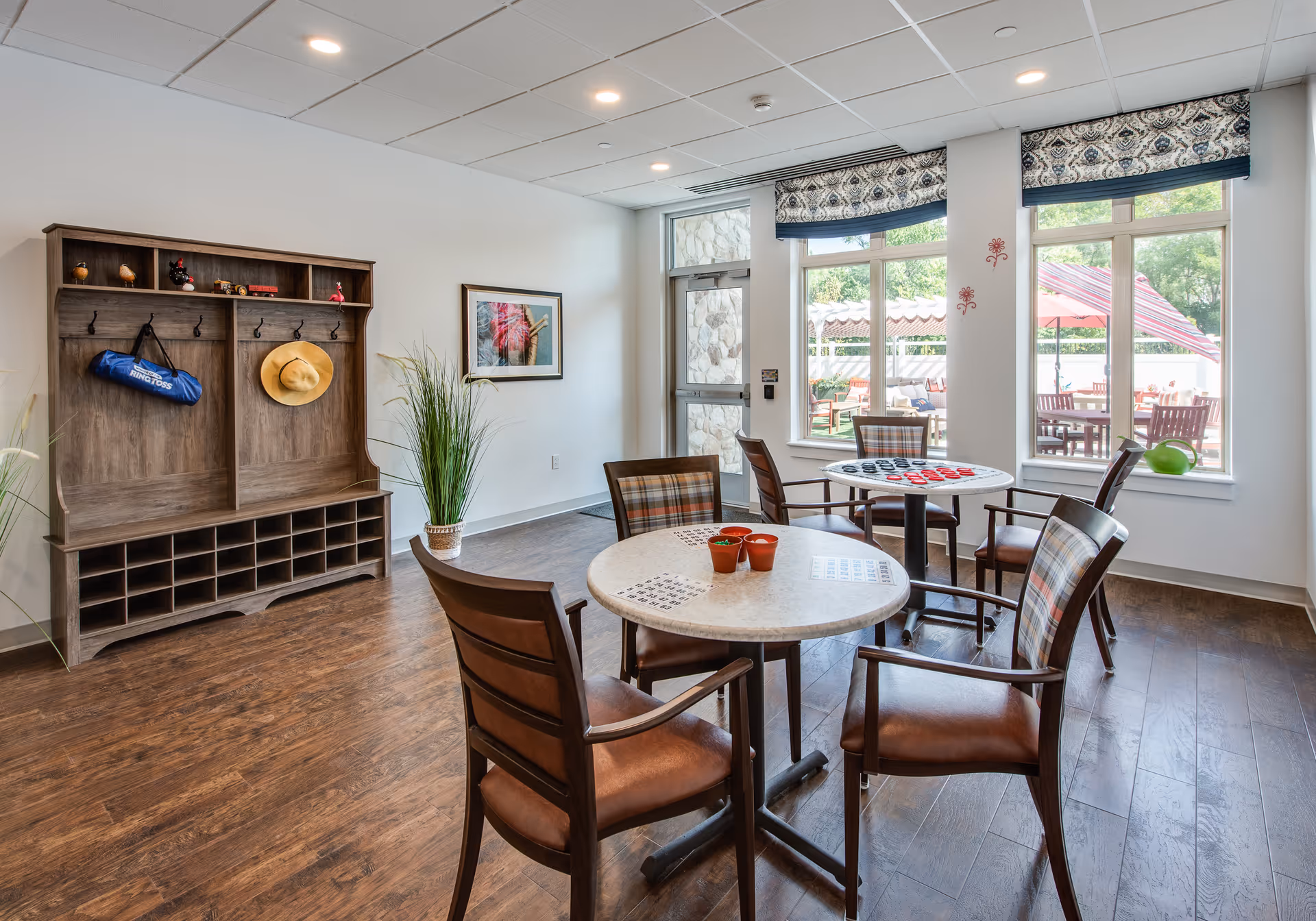 A bright and inviting common area with wooden flooring, featuring two round tables with chairs around them. One table has small potted plants, and the other has a checkers game set up. Large windows with patterned valances let in natural light and offer a view of an outdoor patio with tables and umbrellas. A wooden coat rack with cubbies and hooks is against one wall, holding a hat and a bag. A framed picture and a tall potted plant decorate the room.